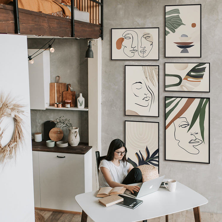 a woman working from home in a tiny studio next to a collection of wall framed mounted beside her on a grey wall, you can see a bed on top of her kitchen that give that industrial decor vibes
