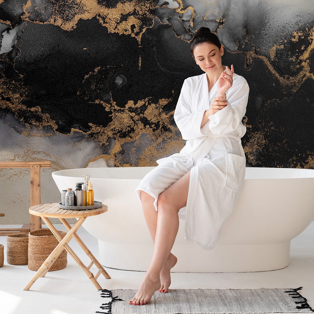 A woman in a white bathrobe sits on the edge of a modern bathtub, applying lotion. Behind her, a black and gold marble-effect wallpaper creates a luxurious backdrop.