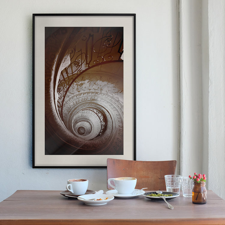 A framed poster of a spiral staircase hangs on a table with two cups of coffee.