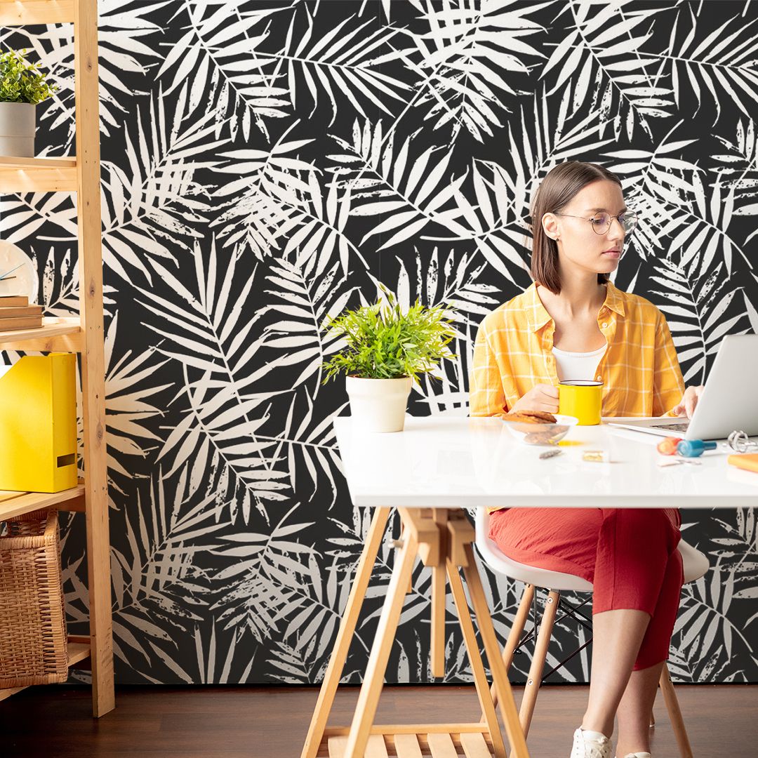 a woman sitting at her home office holding a yellow cup and there's a wallpaper mural with black background and floral motifs in the background